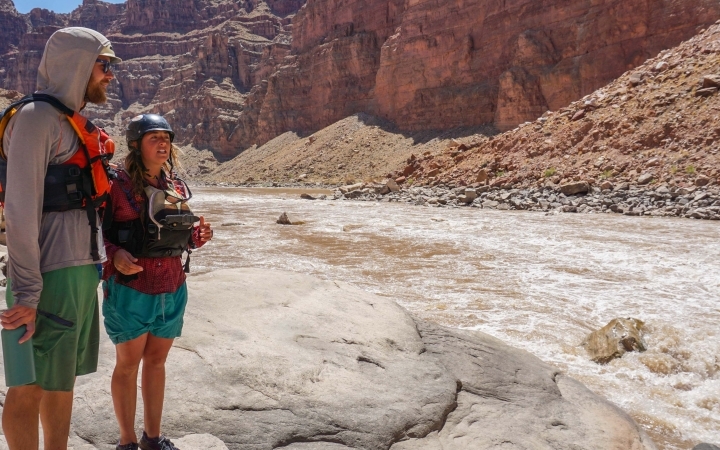 two people wearing life jackers stand on a rock beside a river. There are high canyon walls in the background.